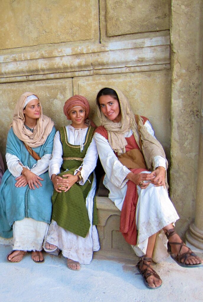 Three women in historical attire sit closely together against a textured wall. Their expressions convey friendship and warmth, highlighting cultural attire.