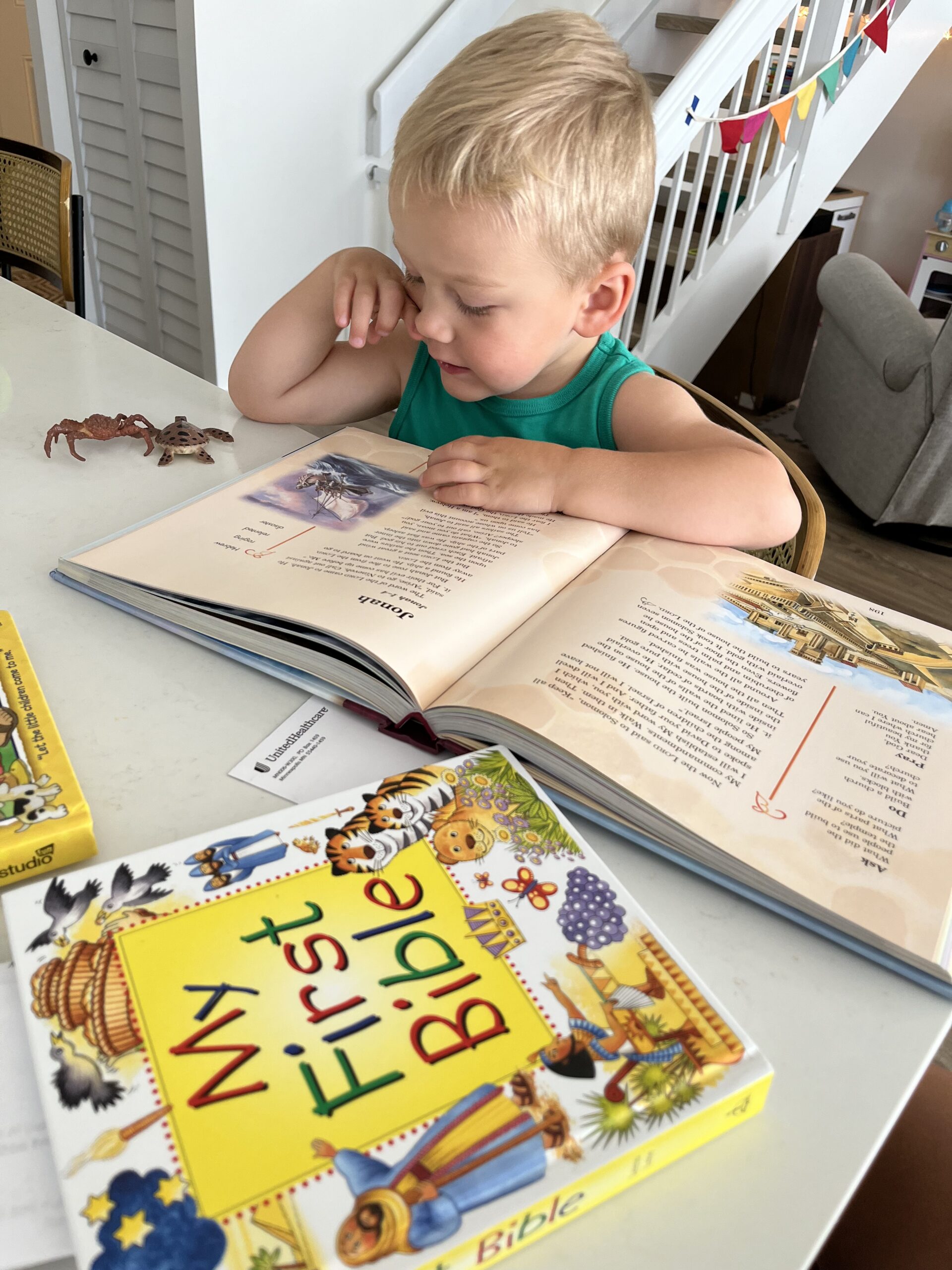 A young boy reads "My First Bible" at a kitchen table, with curiosity on his face. Nearby, colorful toys create a playful atmosphere, enhancing the scene.